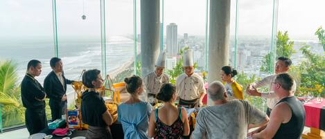 A group of people gathered around a table with a view of the ocean and cityscape through large windows.