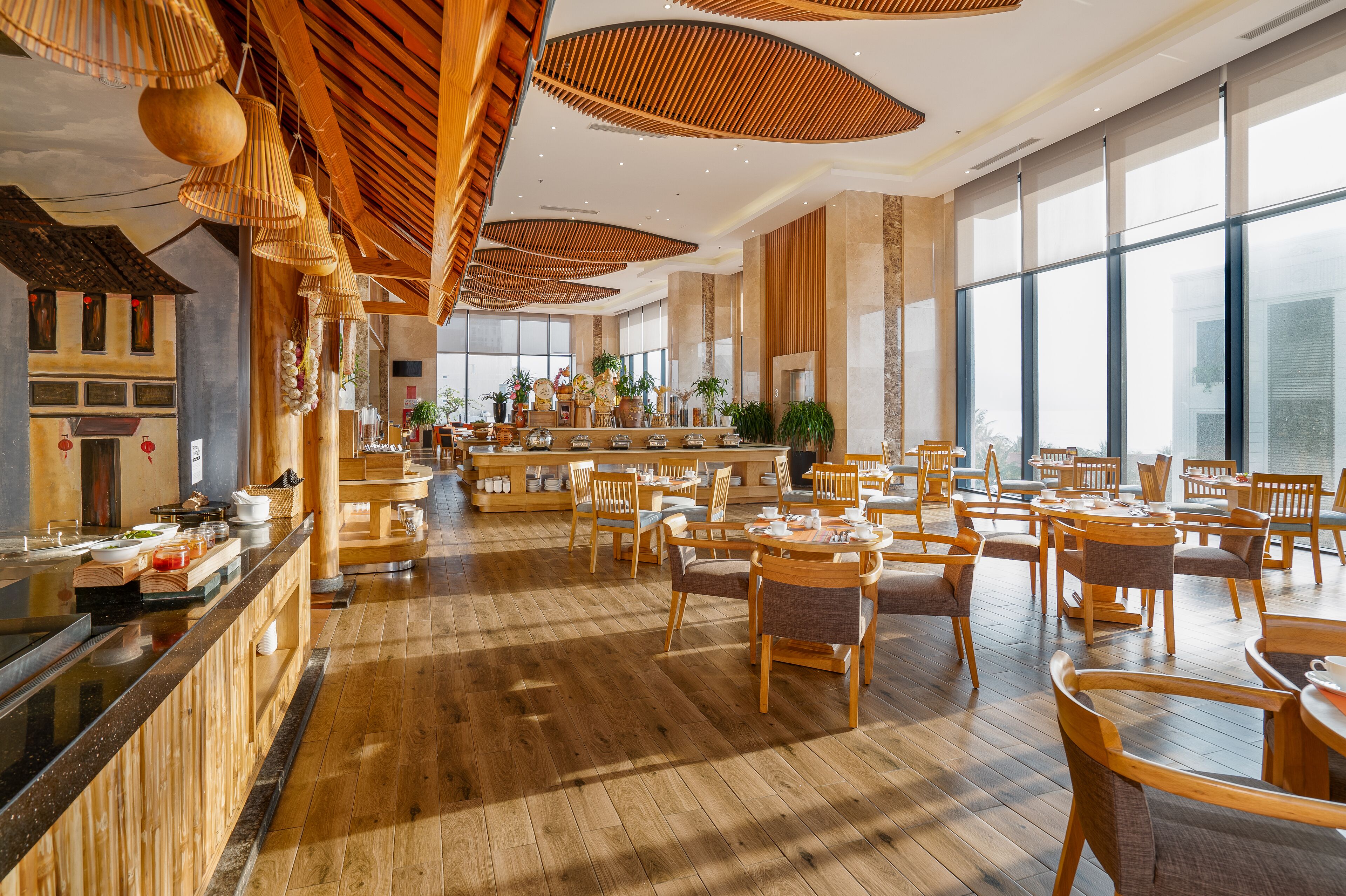 A spacious restaurant with wooden flooring, round tables, and chairs. There are large windows allowing natural light, and a counter with food items.