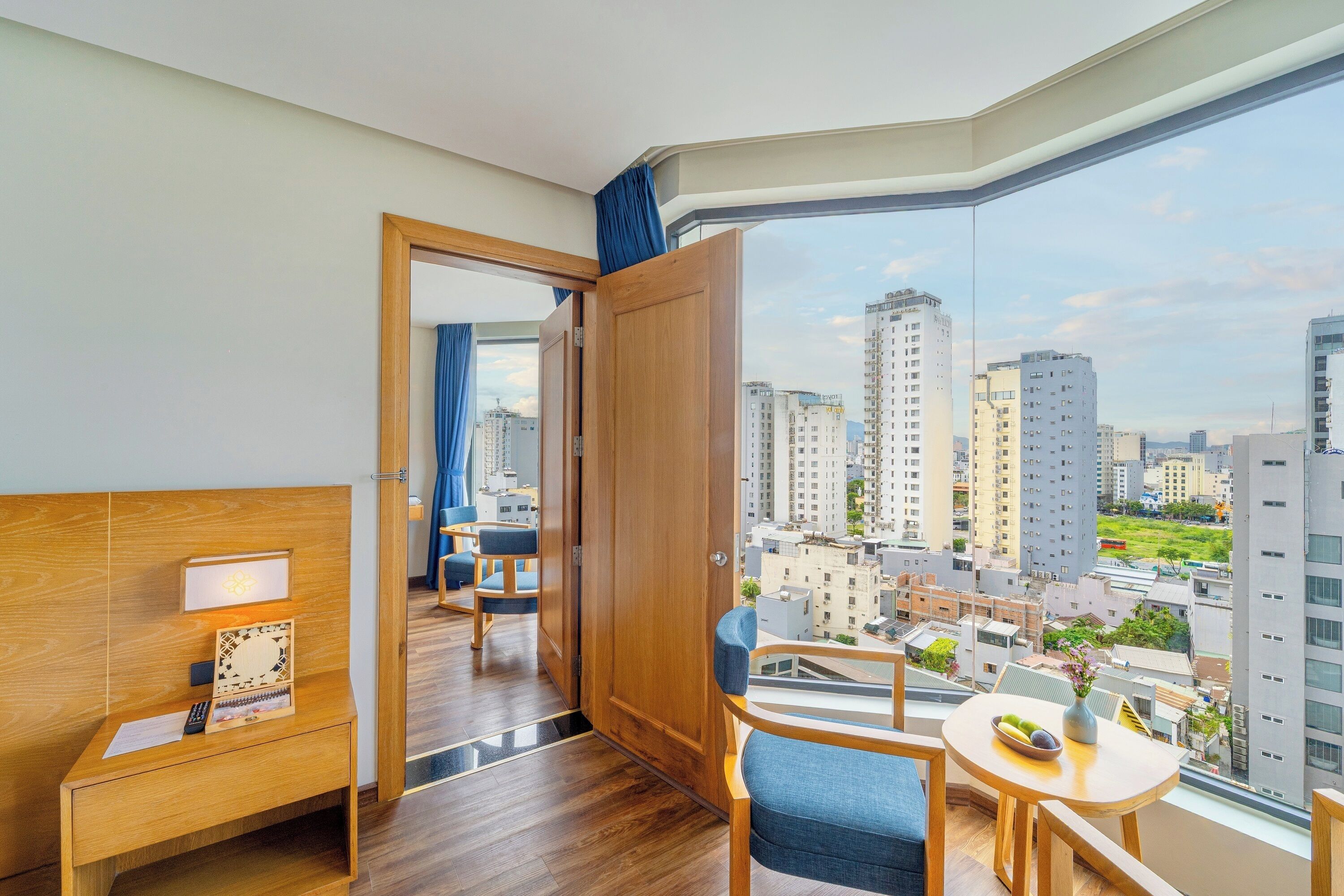 A hotel room with a wooden desk, a small table with fruit, a view of a cityscape, and a large window with curtains.