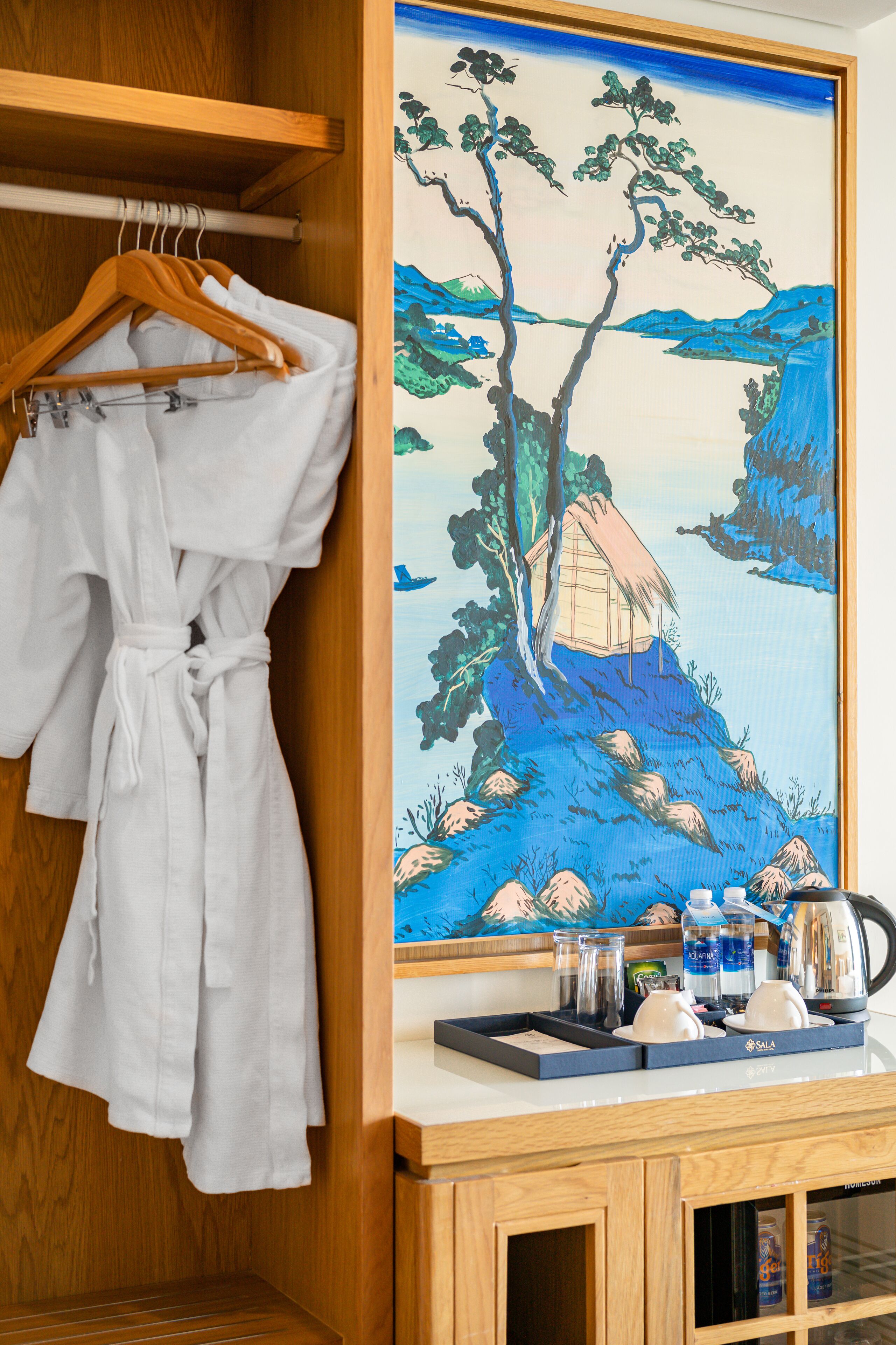 A wooden wardrobe with a white robe hanging, a painting of a landscape, and a minibar with glasses and a kettle.