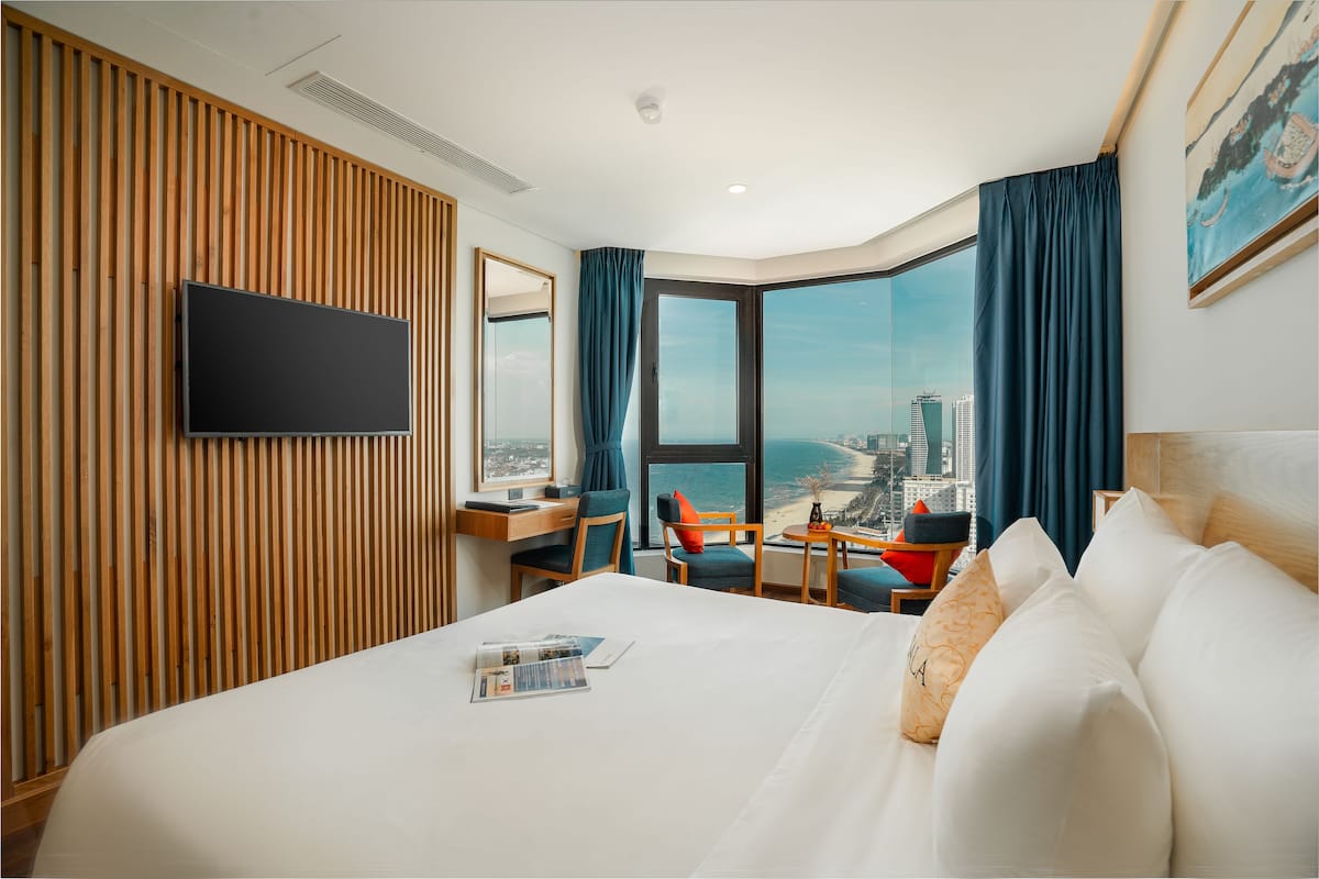 A hotel room with a large bed, a desk, a chair, a TV, and a view of the city and beach.