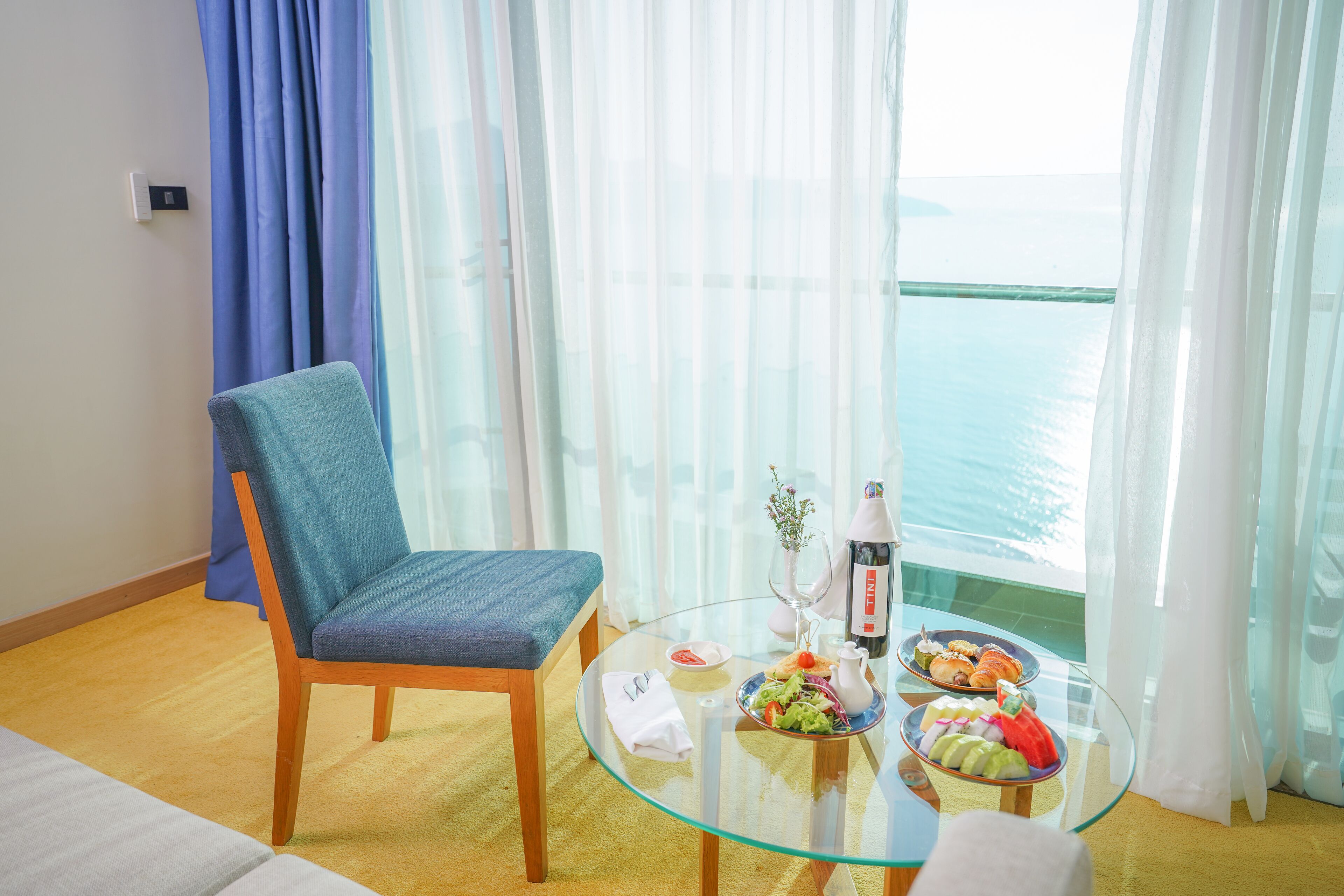 A room with a glass table, a chair, a dining table with food, a bottle, and a view of the sea.