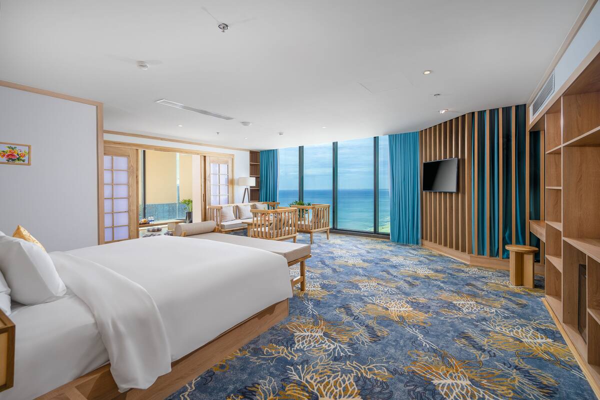 A modern hotel room with a large bed, a seating area, a TV, and a view of the ocean.