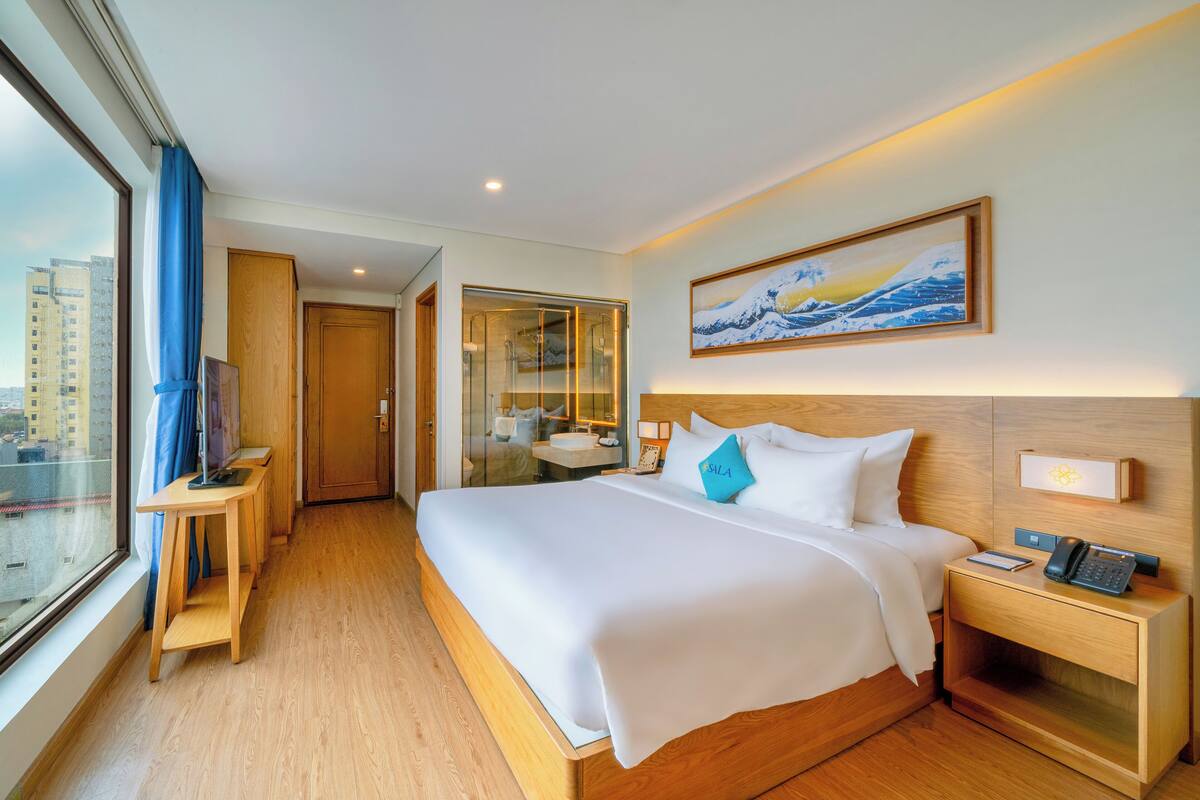 A hotel room with a large bed, a bedside table with a phone, a nightstand, a window with a view of buildings, and a painting on the wall.