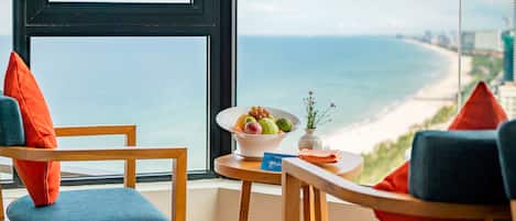 A room with a view of the beach, two chairs with cushions, a small table with a bowl of fruit and a vase with flowers.