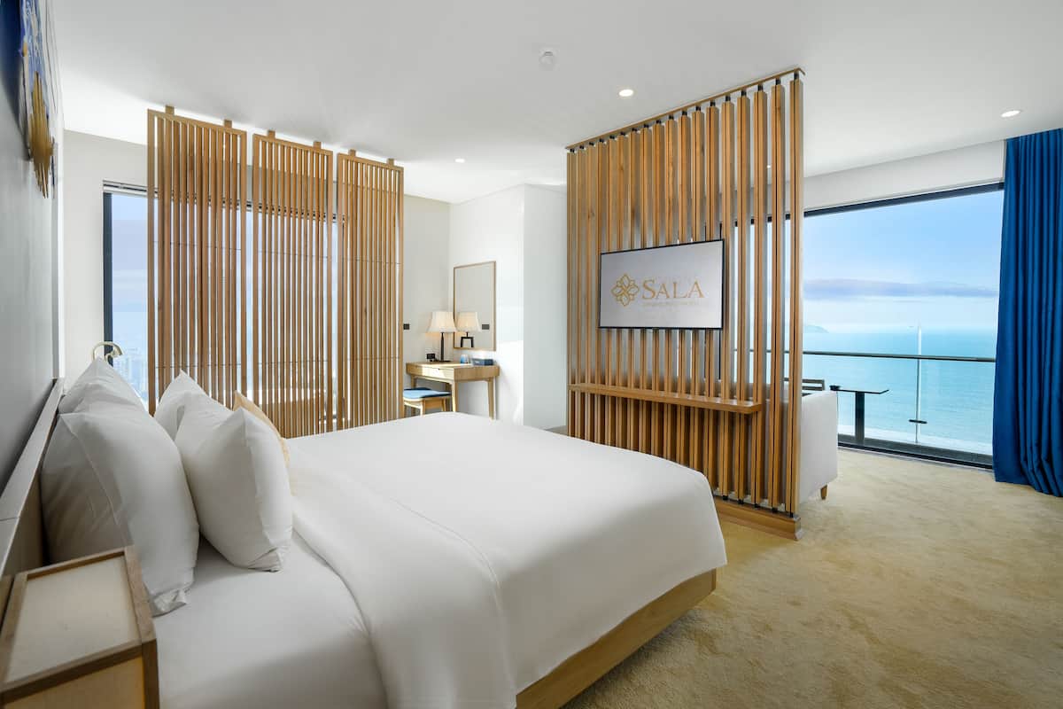 A hotel room with a large bed, a desk, a TV, and a view of the sea.