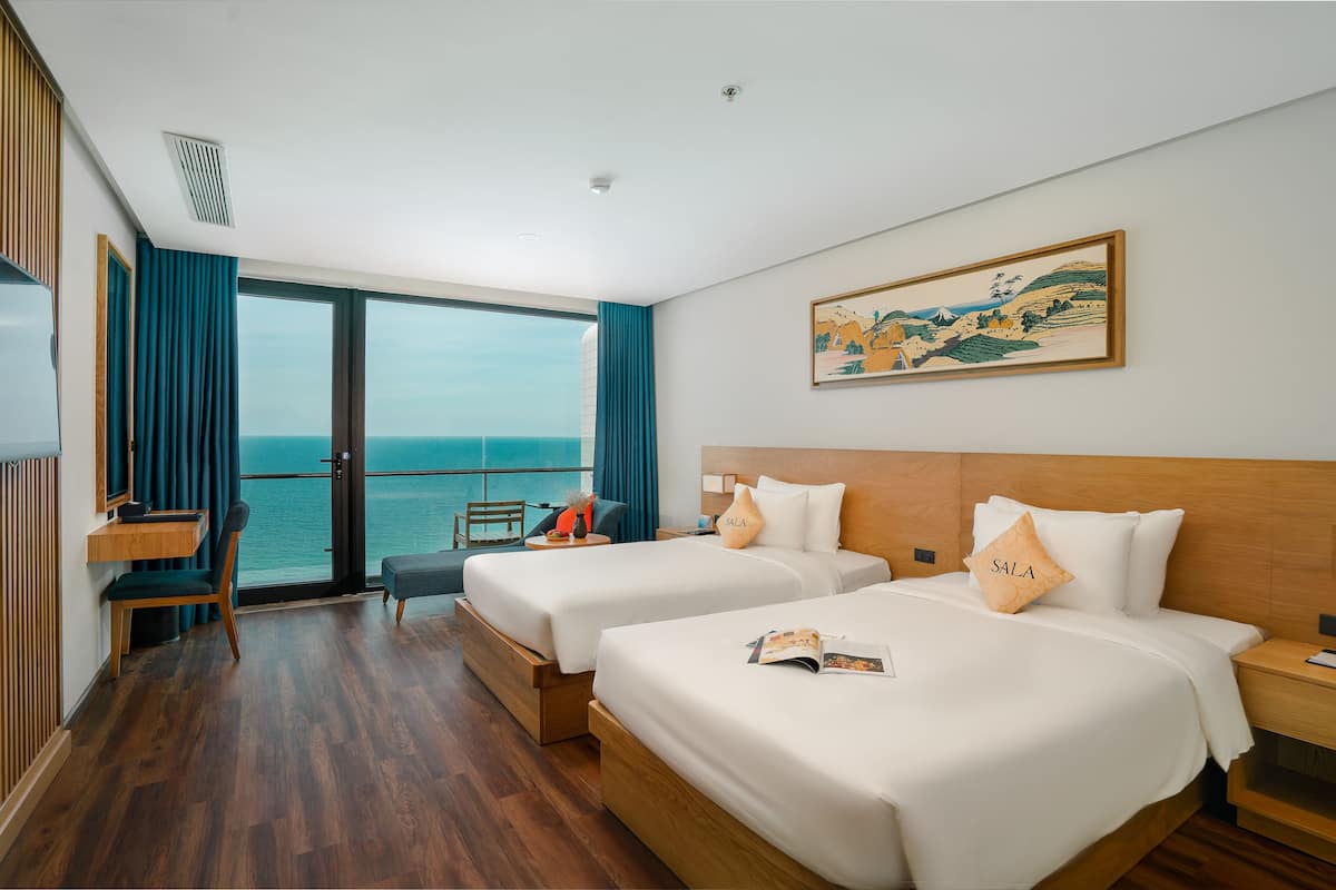 A hotel room with two beds, a desk, a chair, and a large window with a view of the sea.