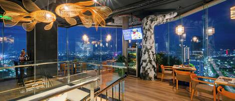 A modern restaurant with a city view, glass railings, and contemporary lighting fixtures.
