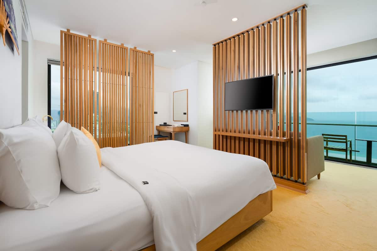 A modern hotel room with a large bed, a desk, a TV, and a balcony with a view of the city.