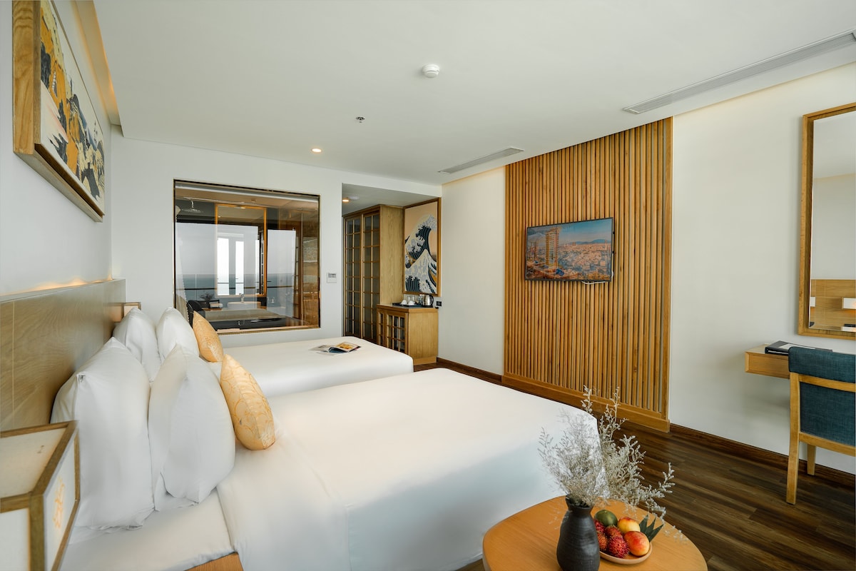 A hotel room with a large bed, a small table with fruit, a wooden panel wall, and a painting.