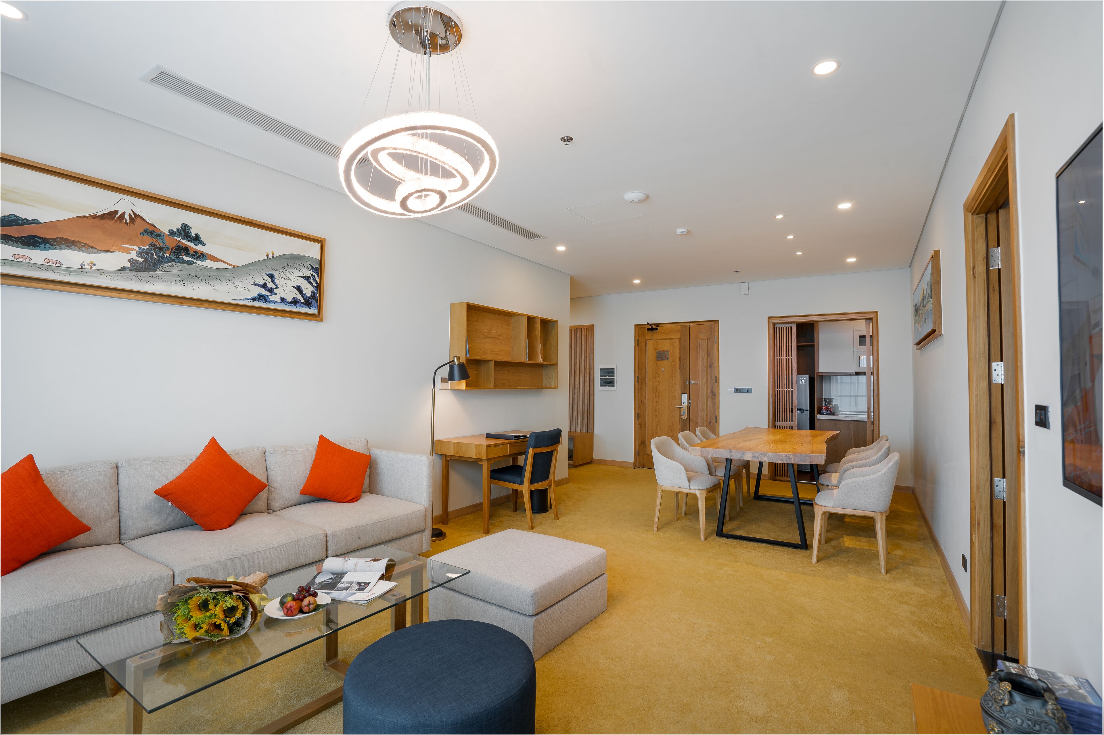 A modern living room with a sofa, ottoman, and glass coffee table. There is a dining area with a table and chairs, and a kitchen visible through an open door.