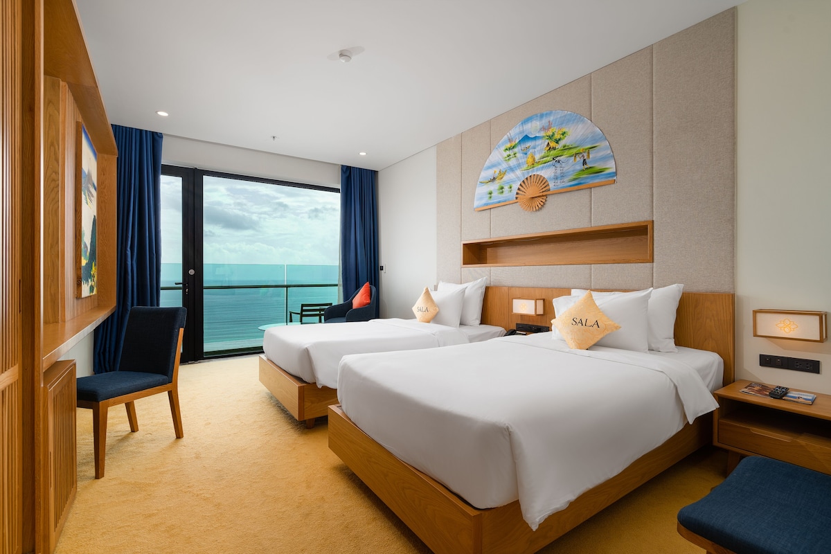 A hotel room with two beds, a desk, a chair, and a view of the sea.