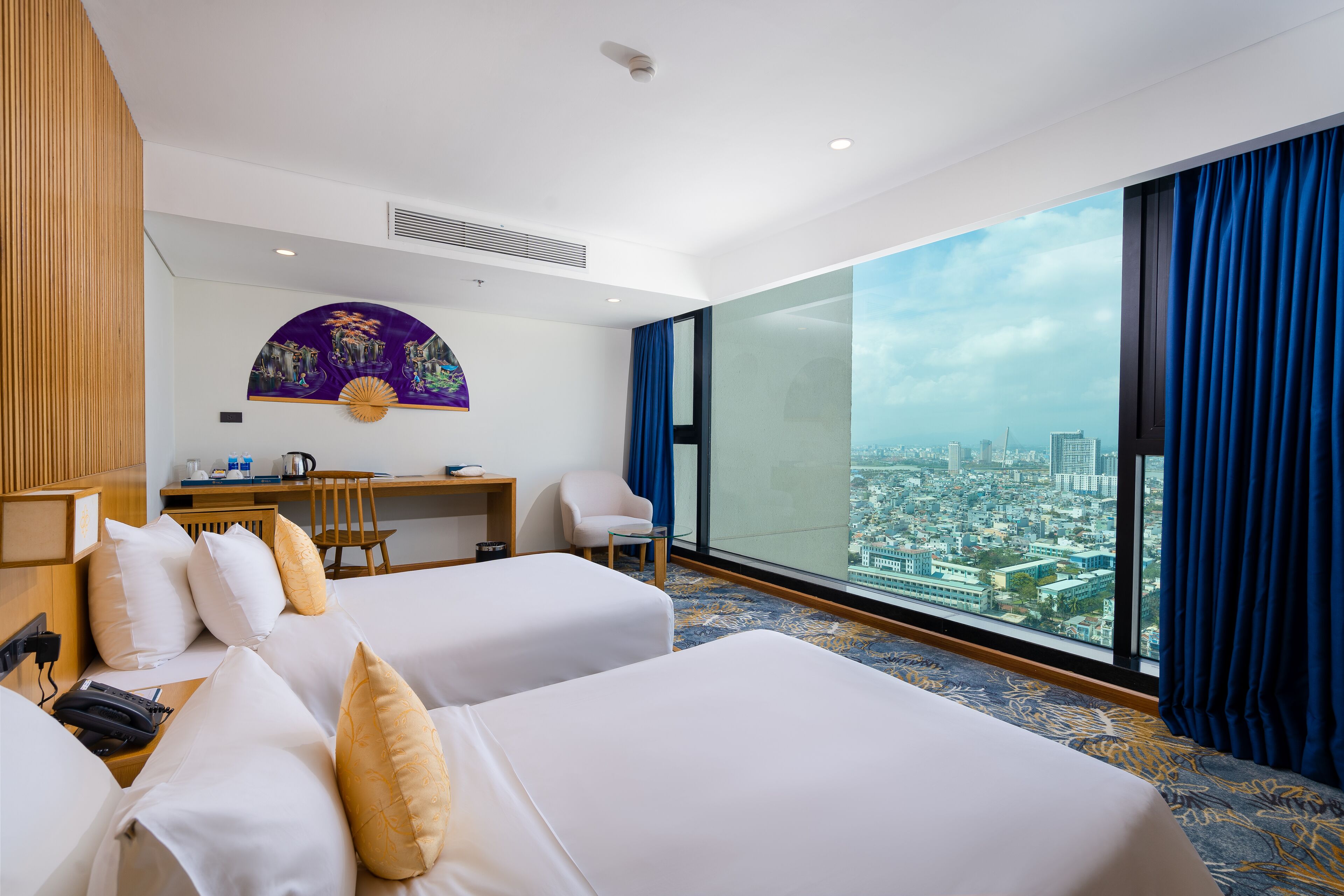 A hotel room with two beds, a desk, a chair, and a large window offering a city view.