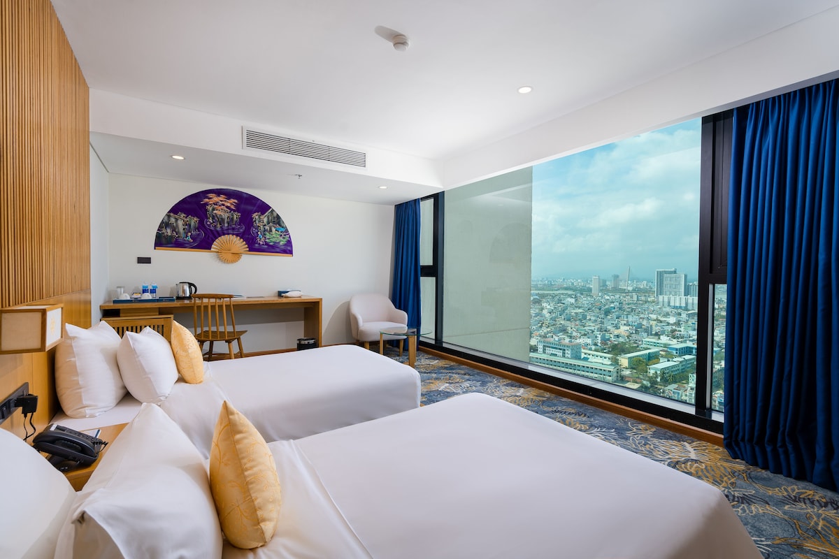 A hotel room with two beds, a desk, a chair, and a large window offering a city view.
