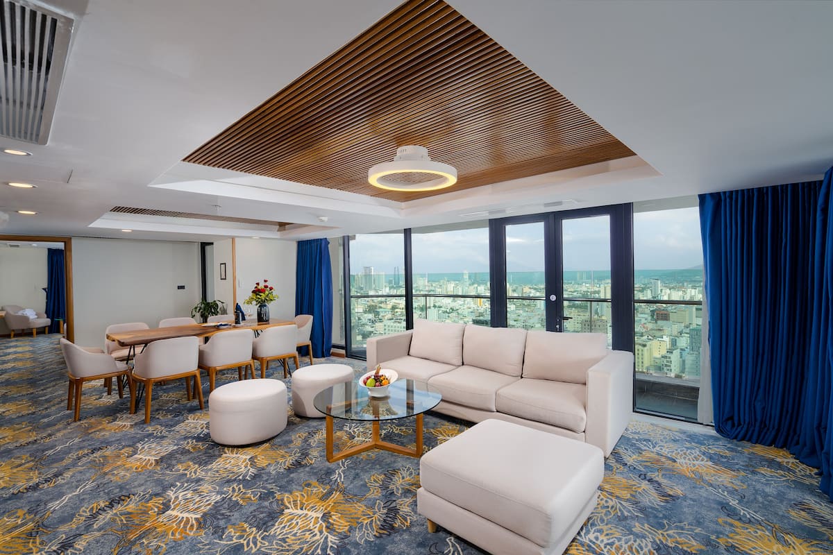 A modern hotel room with a large window offering a city view, a wooden ceiling design, a round glass coffee table, and a beige sofa.