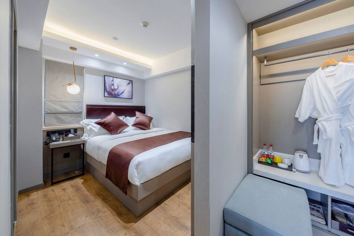 Ein modernes Hotelzimmer mit einem ordentlich bezogenen Bett, einem Nachttisch mit Lampe, einem Kleiderhaken mit Bademantel und einer kleinen Ablagefläche mit Pflegeprodukten.