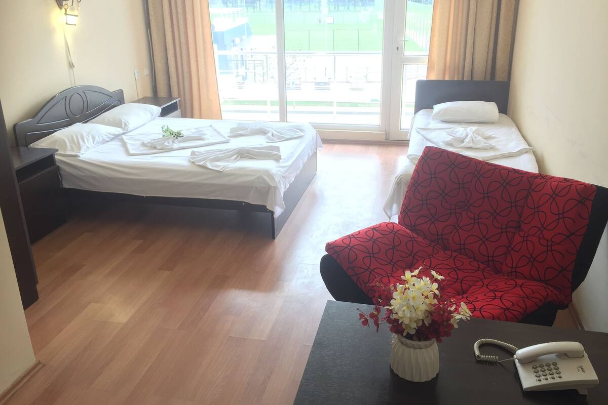 A hotel room with two beds, a red patterned chair, a small table with a vase of flowers, and a telephone.
