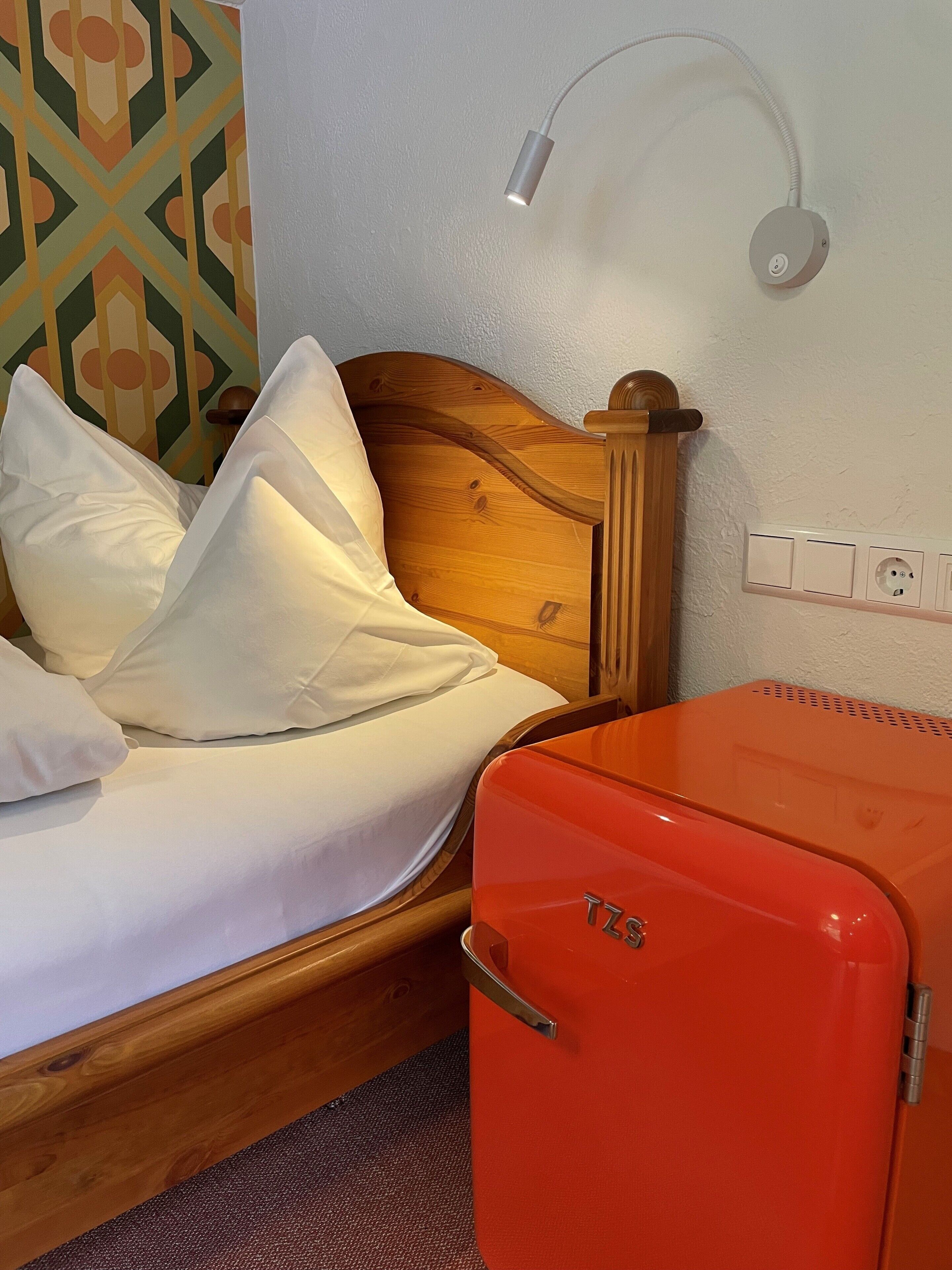A bed with white bedding, a wooden headboard, a wall-mounted lamp, and a red suitcase.