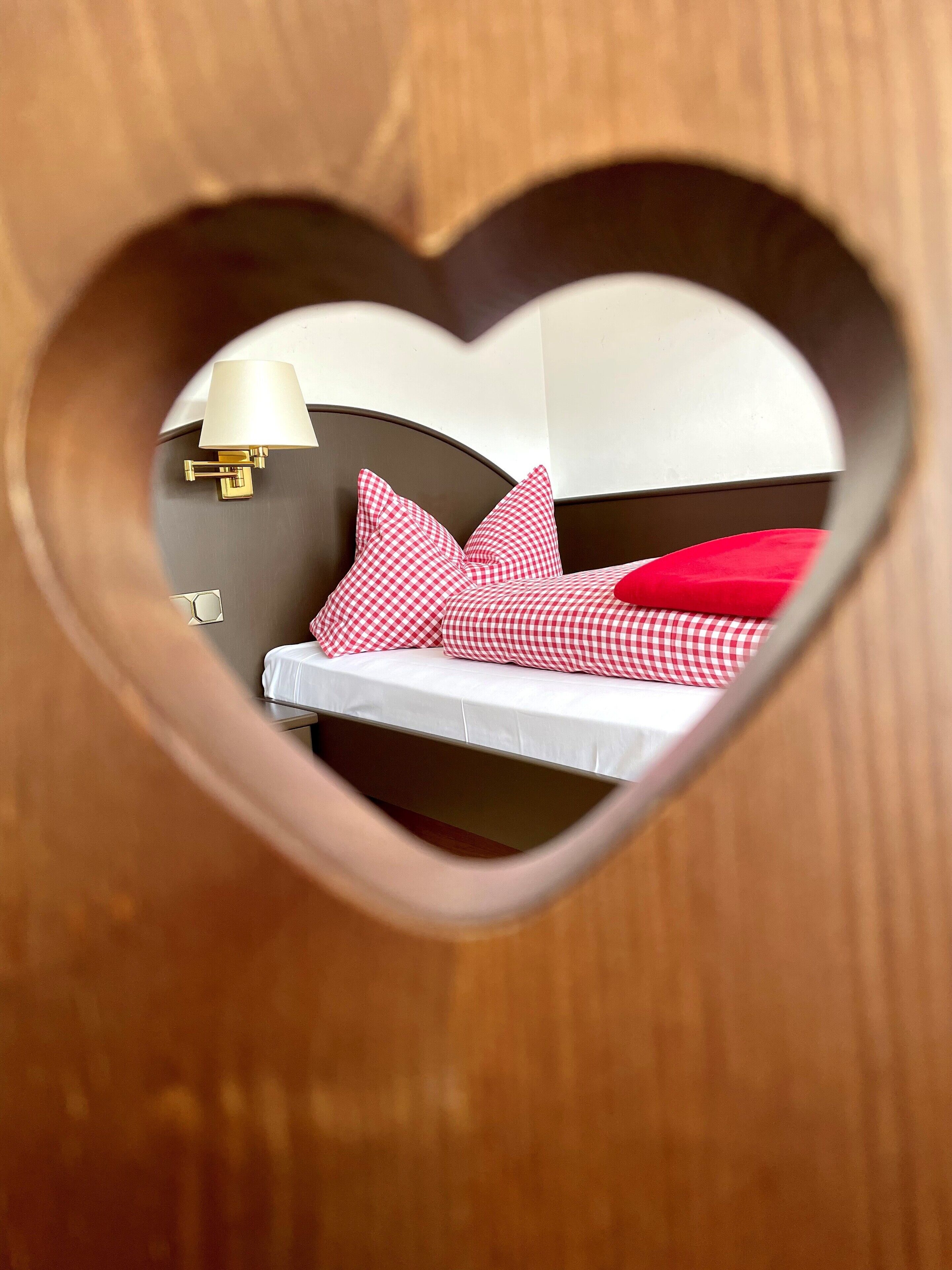 Un’apertura a forma di cuore rivela un letto con copriletto a quadretti bianchi e rossi, una lampada e un punto luce a parete.