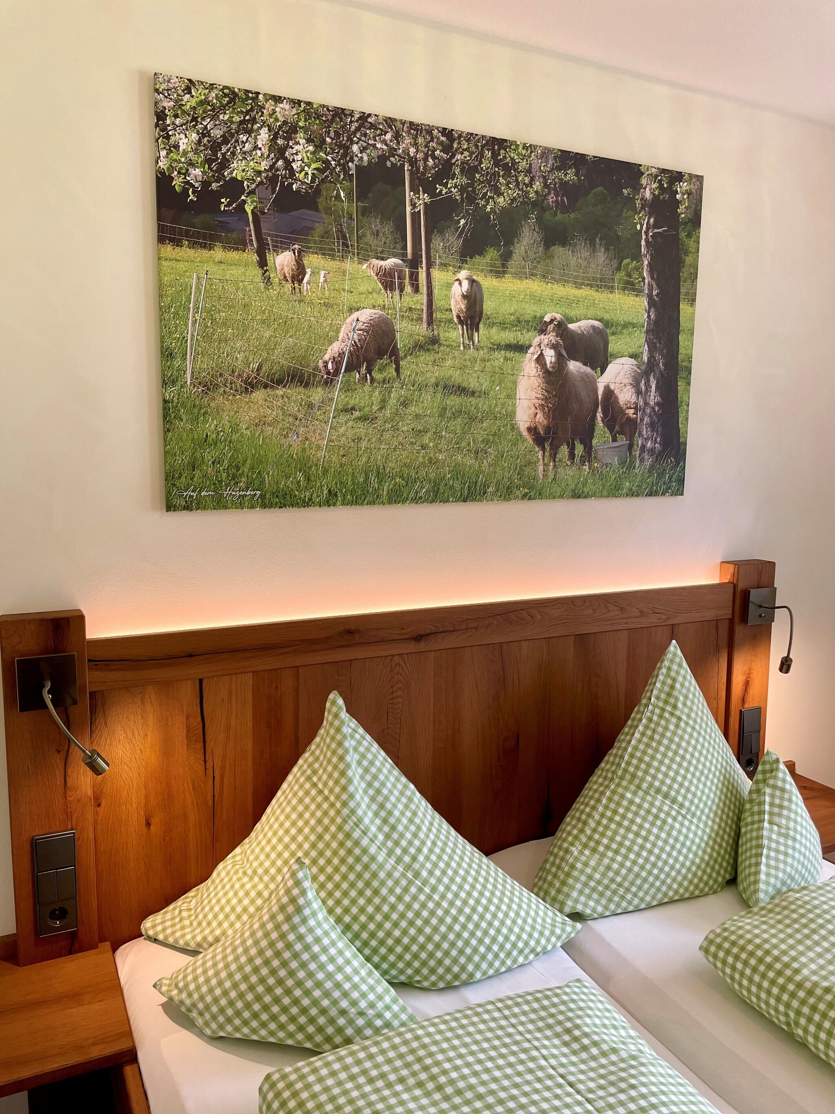 Een slaapkamer met een houten hoofdbord, groene geruit kussens en een aan de muur hangend schilderij van schapen op een veld.