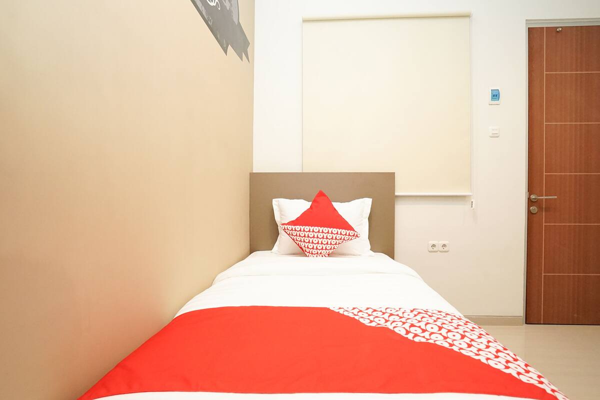A neatly made bed with a red and white bedspread and pillows, a wooden door, and a white wall with a small blue sign.