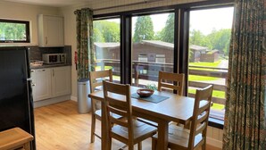 Eine moderne Küche mit weißen Schränken, einem schwarzen Kühlschrank und einer Mikrowelle. Ein Esstisch mit vier Holzstühlen und einer Schüssel steht daneben. Große Fenster mit gemusterten Vorhängen bieten Blick ins Freie.