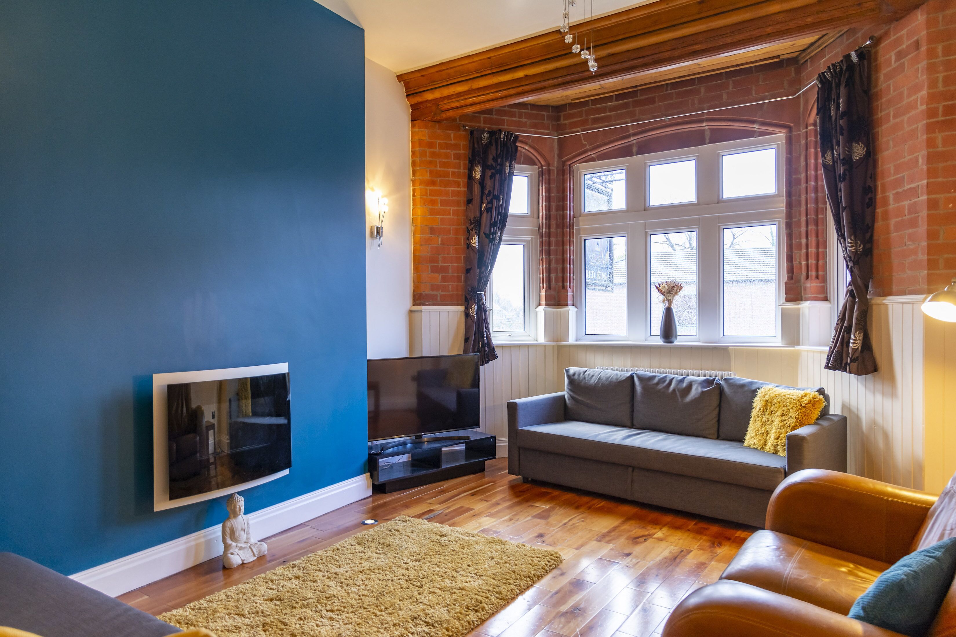 Una sala de estar con pared azul, piso de madera, sofá gris, televisor y ventana con cortinas.