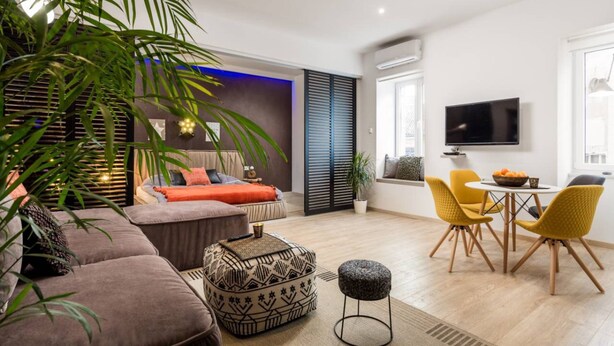 A modern living room with a sofa, ottoman, and yellow chairs. There is a TV mounted on the wall and a window with blinds. - Apartments Kappa (Rijeka)