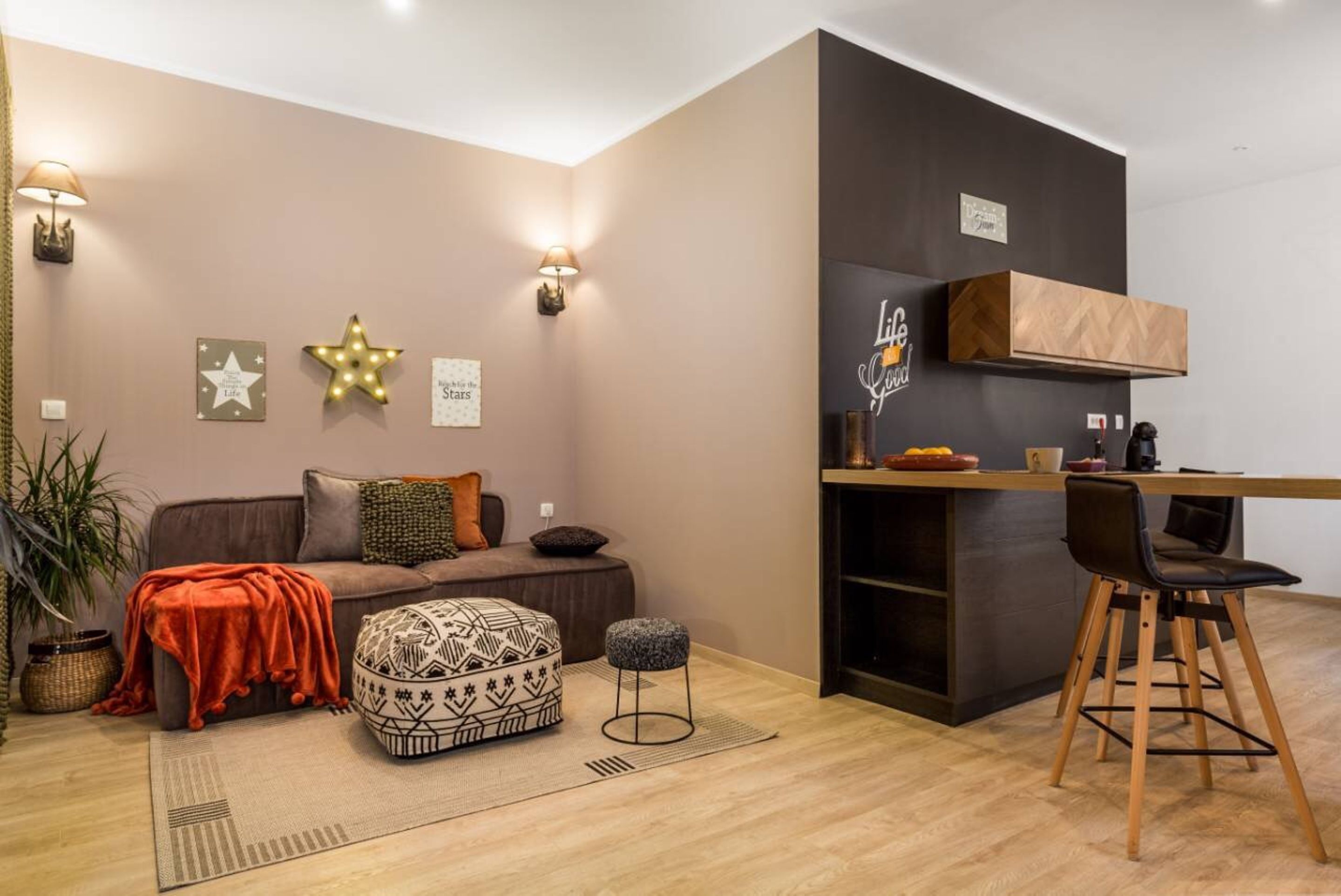 A modern living room with a sofa, ottoman, and a small table with chairs. There is a kitchen area with a countertop and cabinets.