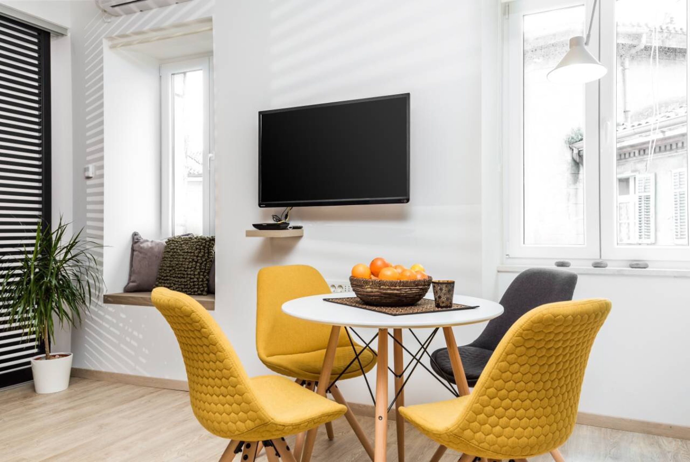 A modern living room with a flat-screen TV mounted on the wall, a round table with a fruit bowl, and yellow chairs.