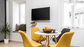 A modern living room with a flat-screen TV mounted on the wall, a round table with a fruit bowl, and yellow chairs.