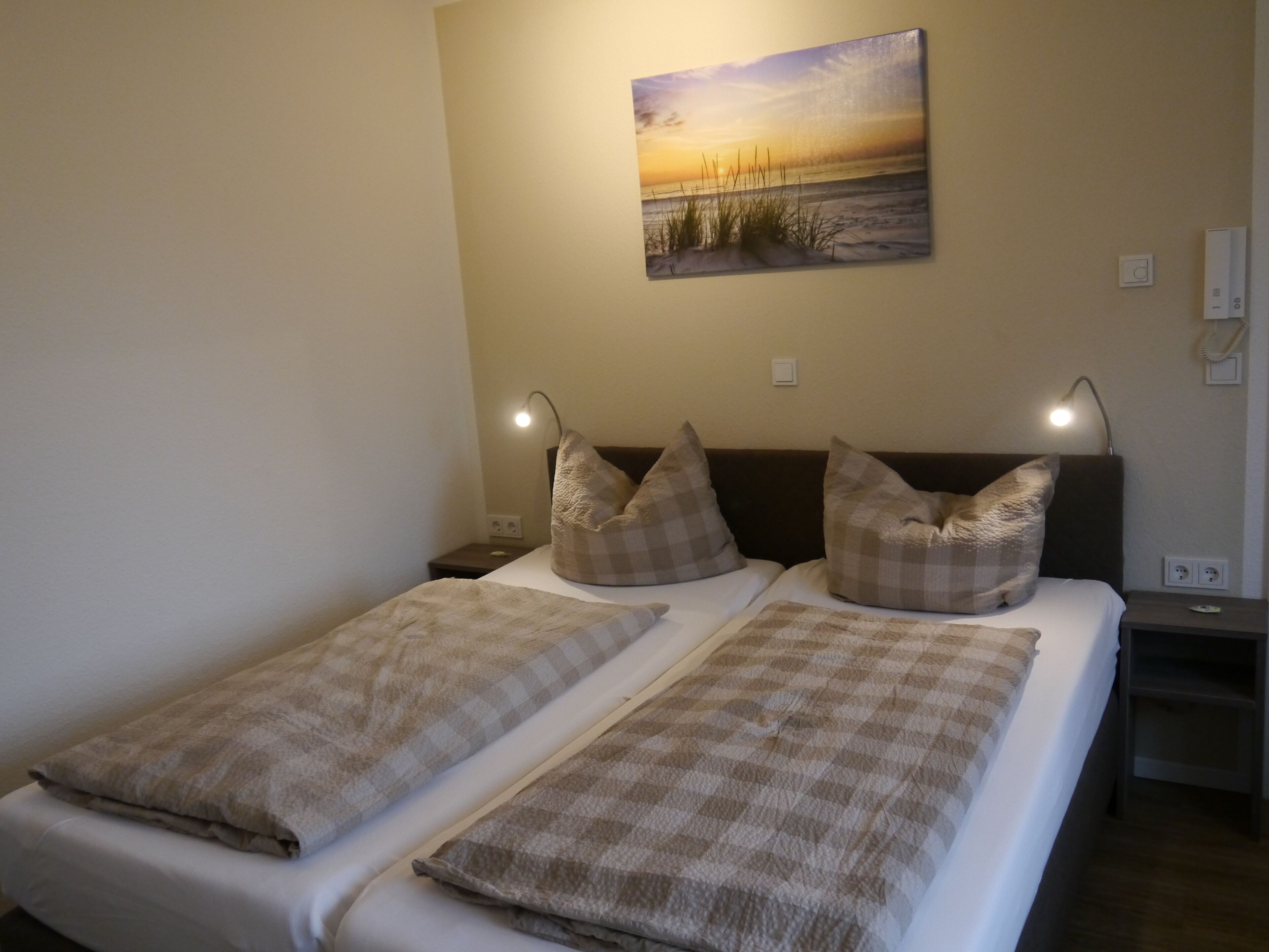 A double bed with checkered bedding, two bedside lamps, a nightstand, and a wall-mounted painting of a sunset over water.
