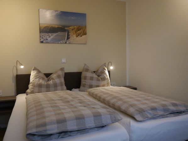 A double bed with checkered bedding, two bedside lamps, and a painting of a beach scene on the wall. - Hotel Haus Wittwer (Emden)