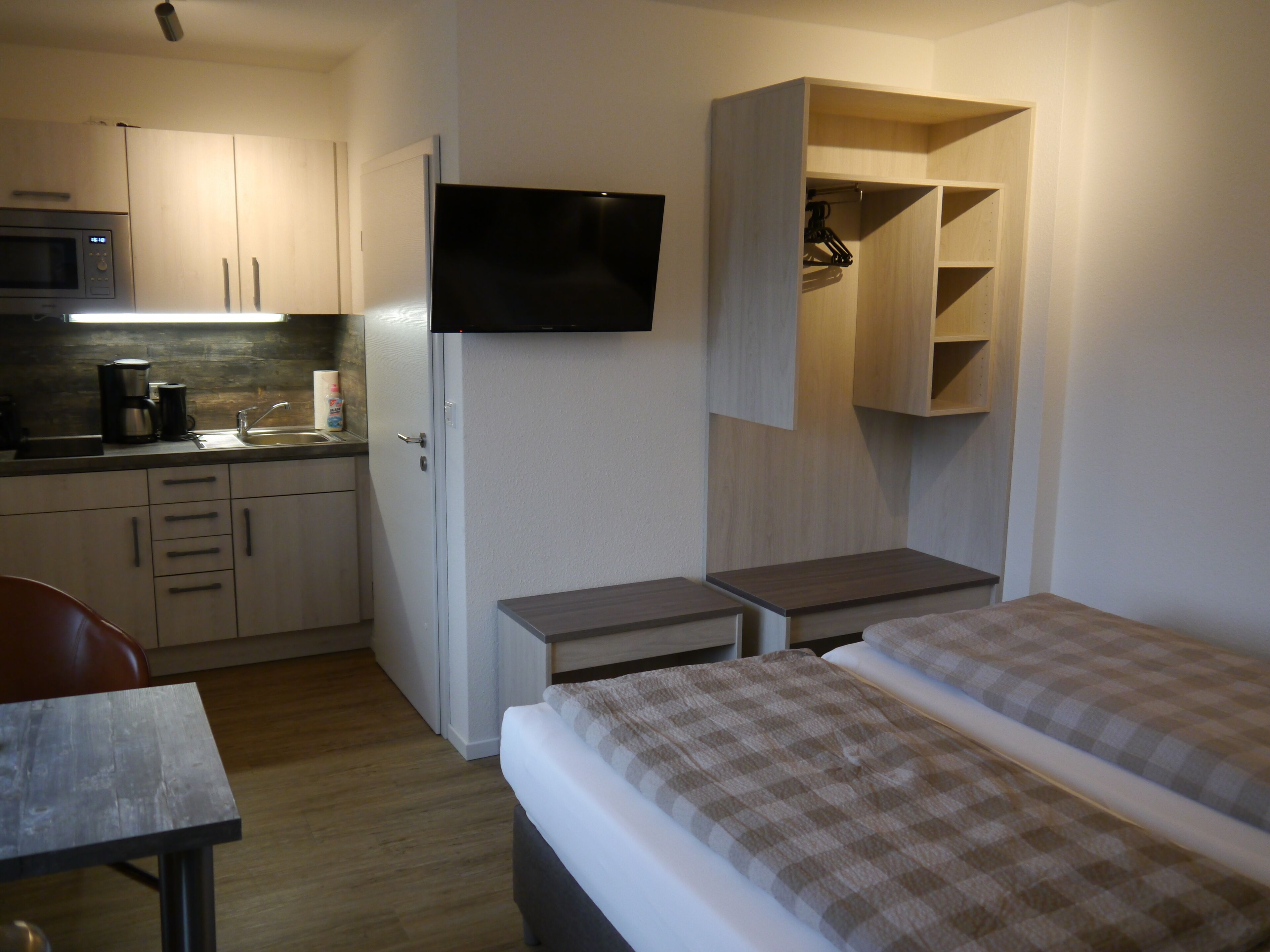 A compact hotel room with a kitchenette, a bed with a checkered duvet, a TV mounted on the wall, and a small table.