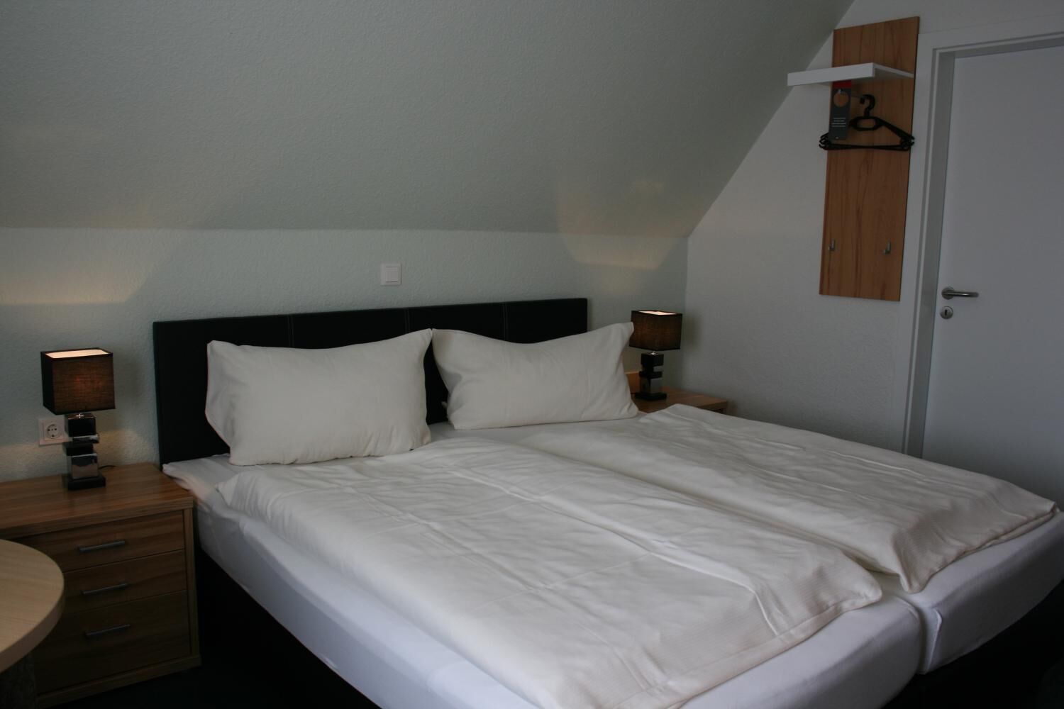 Ein Einzelbett mit weißer Bettwäsche, zwei Nachttischlampen, ein Nachttisch aus Holz und ein kleines Holzschränkchen über der Tür.