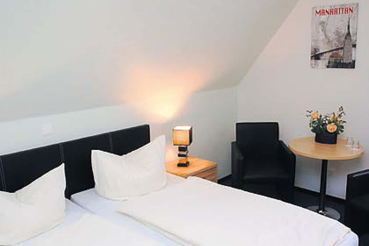 A hotel room with a bed, a nightstand with a lamp, a chair, a small table with a flower vase, and a poster on the wall.