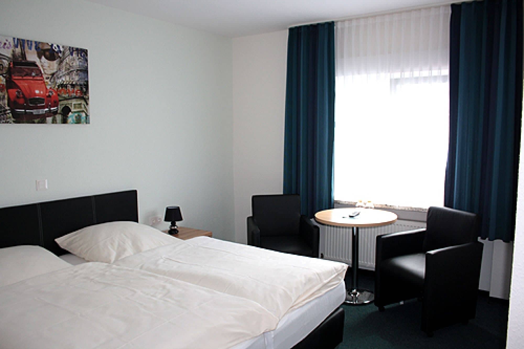 Une chambre d’hôtel avec un lit, deux chaises, une petite table ronde et une fenêtre avec des rideaux.