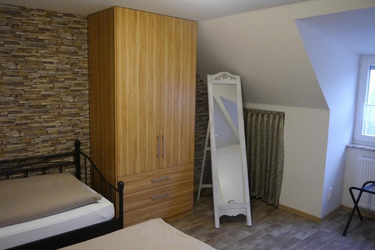 A bedroom with a bed, a wooden wardrobe, a full-length mirror, a radiator, and a window.