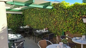 Un patio al aire libre con mesas y sillas, un muro cubierto de enredaderas verdes y un suelo de concreto.