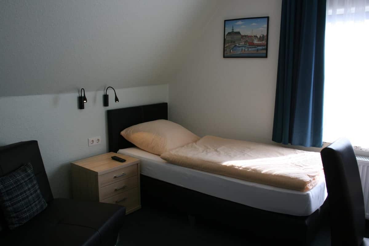 A hotel room with a single bed, a bedside table with a lamp, a desk with a chair, and a painting on the wall.