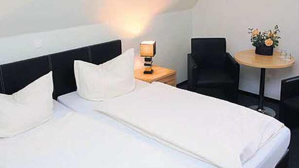 Habitación de hotel con cama, mesita de noche con lámpara, una silla, una pequeña mesa con un florero y un cartel en la pared.