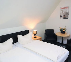 A hotel room with a bed, a nightstand with a lamp, a chair, a small table with a vase of flowers, and a poster on the wall. - Hotel Haus Wittwer (Emden)