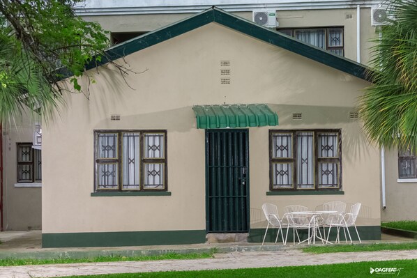 Un edificio con tejado y puerta verdes, y dos ventanas con rejas decorativas. Hay una pequeña mesa con sillas afuera.