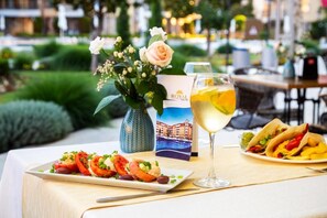 A dining table with a plate of food, a glass of white wine, a vase with flowers, and a brochure. - Aparthotel Royal Sun (Sunny Beach)