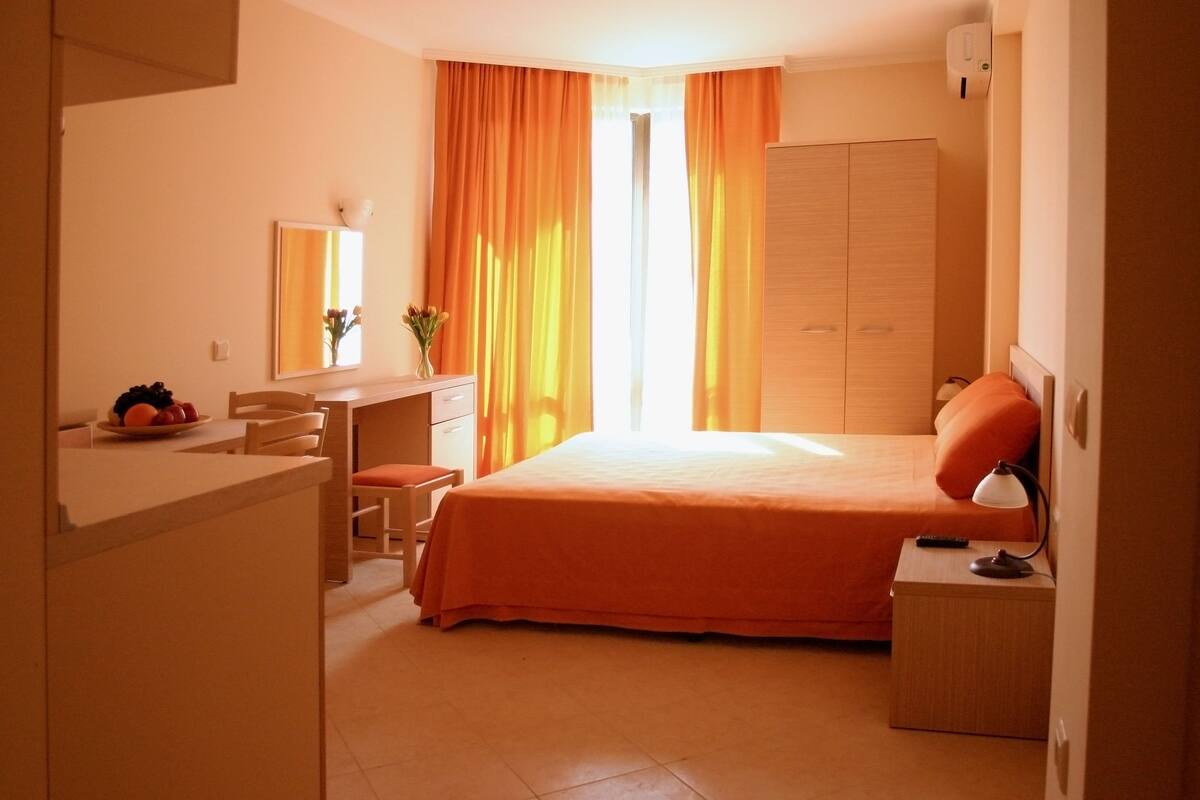 A hotel room with a bed, a desk, a chair, a wardrobe, and a small table with a lamp.