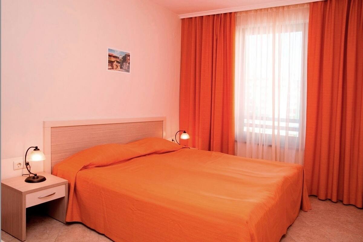 A hotel room with a single bed, two bedside lamps, a nightstand, and a window with orange curtains.