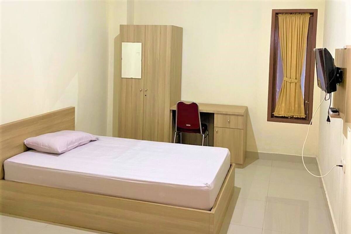 A single bed with a wooden frame, a desk with a chair, a wardrobe, and a television mounted on the wall.