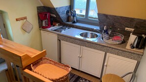 A kitchen with a sink, stove, and a coffee maker. There is a wicker basket and a chair. - Toplicht - Villa Doris (Koserow)
