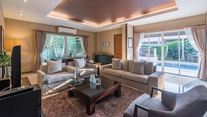 Un salon moderne avec deux canapés, une table basse et un téléviseur. De grandes fenêtres sont agrémentées de rideaux, un luminaire est fixé au plafond, et on peut admirer une piscine depuis l’intérieur.