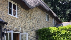A stone house with a thatched roof, a wooden door, and a small patio with a table and chairs. - Thatched Cottage, West Dorset (Dorchester)